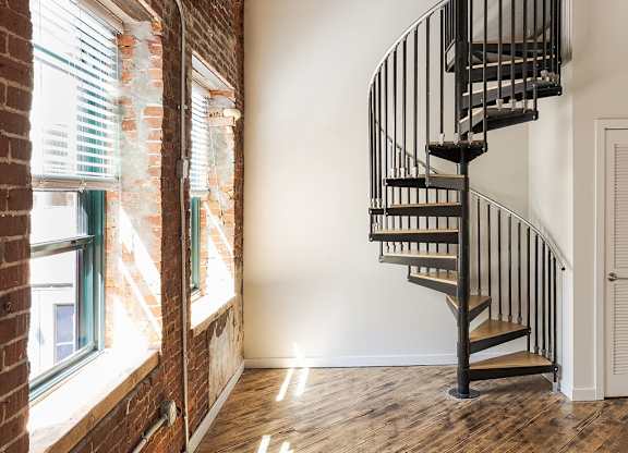 A wooden staircase with a metal railing is in a room with a brick wall and a window.