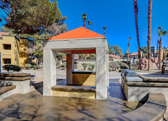 A small gazebo with a red roof is situated in a park-like setting with a bench in front of it.