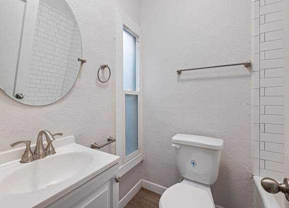 A white toilet with a blue sticker on the lid is in a bathroom with a white sink and mirror at Hillstone Flats Apartments, Los Angeles, CA