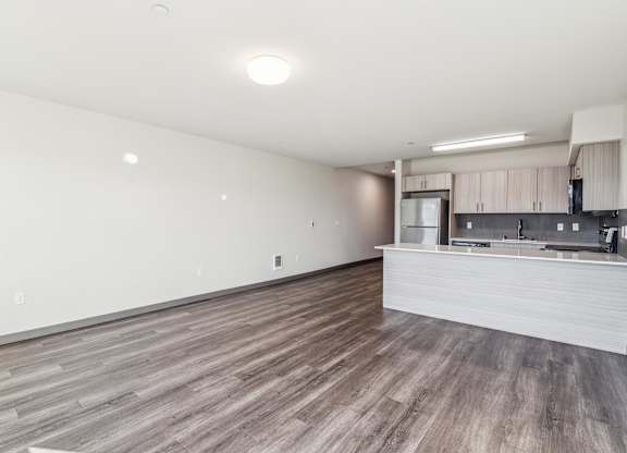 A spacious room with wooden flooring and a kitchen area in the background.