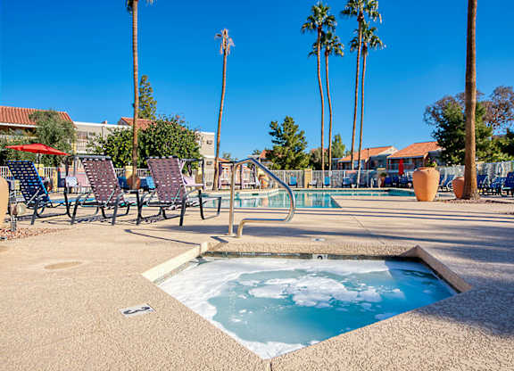 A hot tub sits in the middle of a patio with chairs and palm trees in the background.