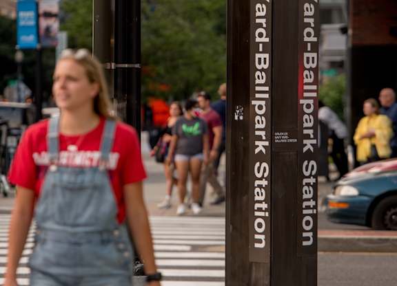 A woman in a red shirt and denim overalls is walking across the street at Navy Yard Ballpark Station.