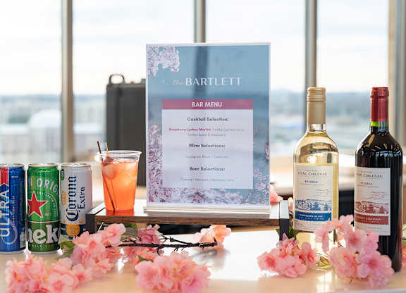 A bar menu is displayed on a table with a can of Corona, a glass of orange juice, and three bottles of wine.