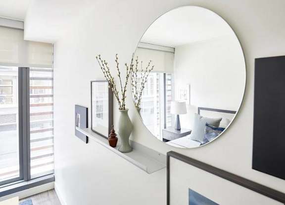 A round mirror hangs on a wall above a shelf with vases and pictures.