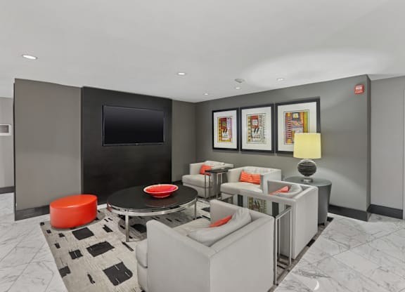 A modern living room with a grey couch, a black and white rug, a red ottoman, a black coffee table, a flat screen TV, and framed pictures on the wall.
