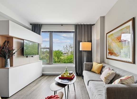 A modern living room with a grey couch, a coffee table, and a large window with a view of the trees outside.