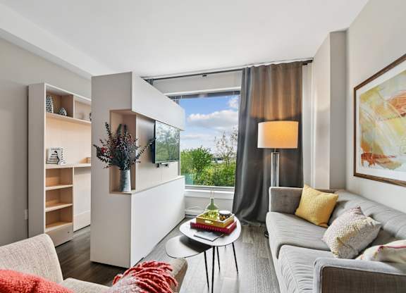 A modern living room with a grey couch, a white coffee table, and a large window with a view of the trees outside.