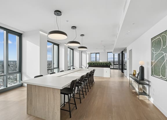 A long white table with black chairs is in a room with a view of the city.