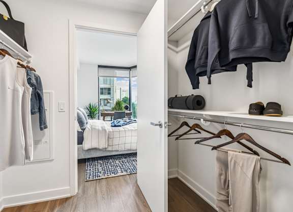 A white closet with clothes hanging and a bedroom in the background.
