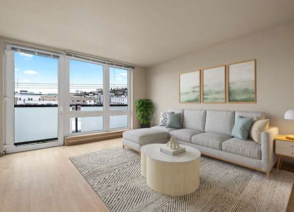 A living room with a grey couch, a coffee table, and a large window overlooking a cityscape.