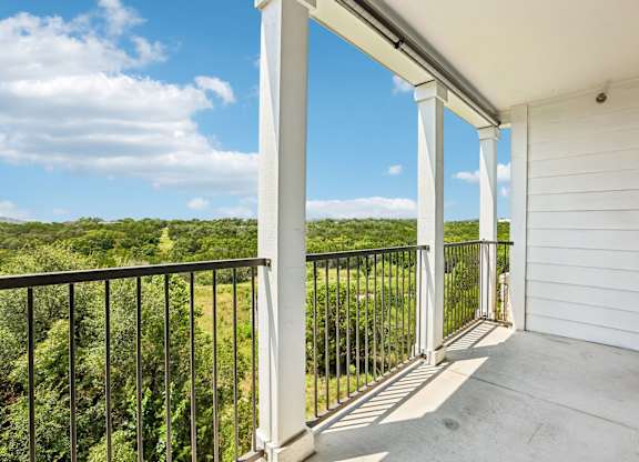A balcony with white columns and a black railing overlooks a green landscape.
