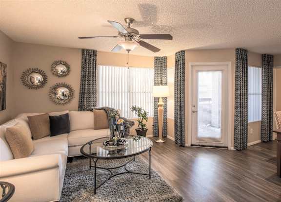 Dog-Friendly Apartments in Ahwatukee, AZ - Pacific Bay Club - Living Room with Wood-Style Plank Flooring, Area Rug, White Sectional Couch, Circular Glass Coffee Table, Dining Area with Table and Chairs, Decorative Art, Standing Lamp, Ceiling Fan, Windows with Curtains, Door with Access to Patio or Balcony.