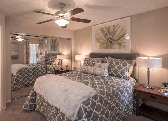 One and Two-Bedroom Apartments in Ahwatukee, AZ - Pacific Bay Club - Bedroom with Mirrored Closet Doors, Carpet Floors, Ceiling Fan, Cozy Bed, Stylish Decor, and Door to Bathroom with Wood-Style Plank