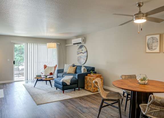Mountain Park, OR, Apartments For Rent - MiLO - Living Room With Wood-Style Flooring, Area Rug, Sectional Couch, Side Table, Floor Lamp, Dining Area, Ceiling Fan, And Sliding Glass Door With Access to Balcony.