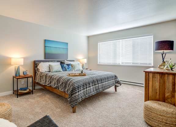 One-Bedroom Apartments in Mountain Park, OR - MiLO at Mountain Park - Bedroom with Carpeting, Large Window, Wooden Dresser, and 2 Nighstands with Lamps