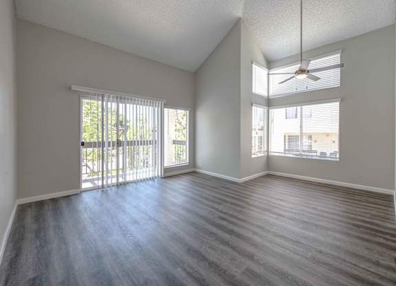 One Bedroom Apartments in San Clemente, CA - Rancho Del Mar - Empty Living Room Area with Wood-Style Flooring, Vaulted Ceilings, Ceiling Fan, Large Windows, and Glass Sliding Door to Private Balcony.