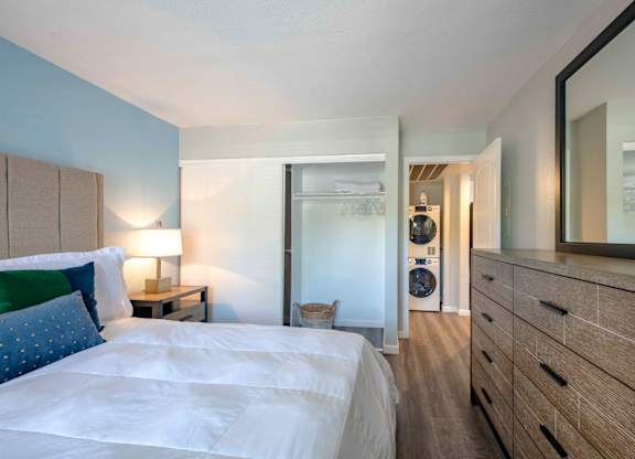 Two Apartments in San Clemente, CA - Rancho Del Mar - Bedroom with Wood-Style Flooring, Nightstand with Lamp, Dresser, Mirror, Spacious Closet with Sliding Door, and View of Washer and Dryer.