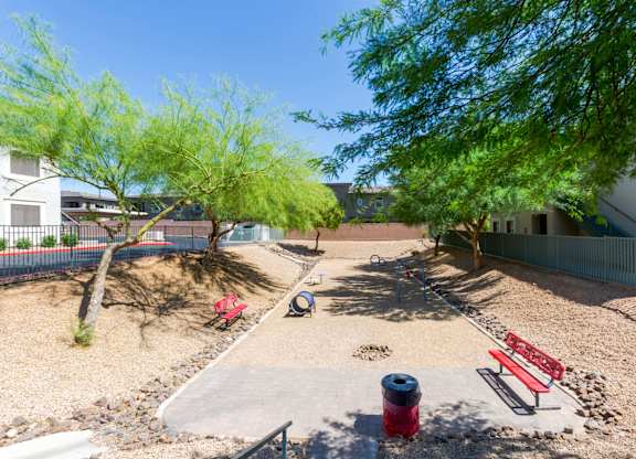 Three Bedroom Apartments in Scottsdale, AZ - The Sycamore at Scottsdale - Outdoor Dog Park with Partial Stone Tile Area, Red Benches, Red Trash Can, Long Dirt Covered Area with Dog Play Structure and Agility Equipment, and Surrounding Gravel, Trees, and Fencing.