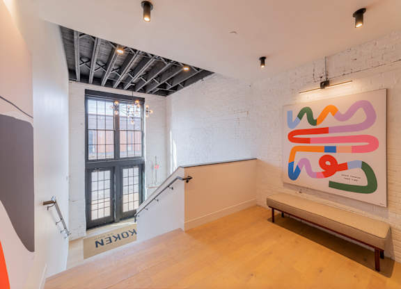 A large abstract painting hangs on a white brick wall in a spacious room with wooden floors and a metal staircase.