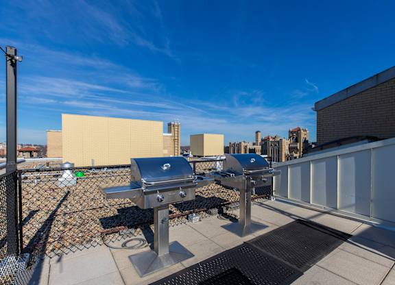 A grill is on a patio with a white fence and a building in the background.
