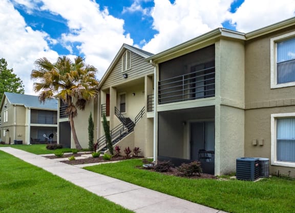Exterior view of a modern apartment building with beige stucco walls, screened-in balconies, and landscaped walkways. The community features lush green lawns, palm trees, and a bright blue sky with scattered clouds