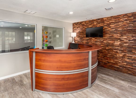 Reception area with a wooden desk and a wall of stacked wood panels. A window behind the desk leads to a gym where colorful medicine balls are visible through the glass. A TV is mounted on the wall above the desk