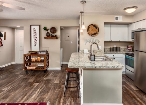 Spacious open-concept kitchen with granite countertops, stainless steel appliances, and white cabinetry. The kitchen features a small island with a stool, and the wall above displays a 'gather' sign. The living area has decorative accents, including framed artwork, a wooden shelf, and a rolling cart