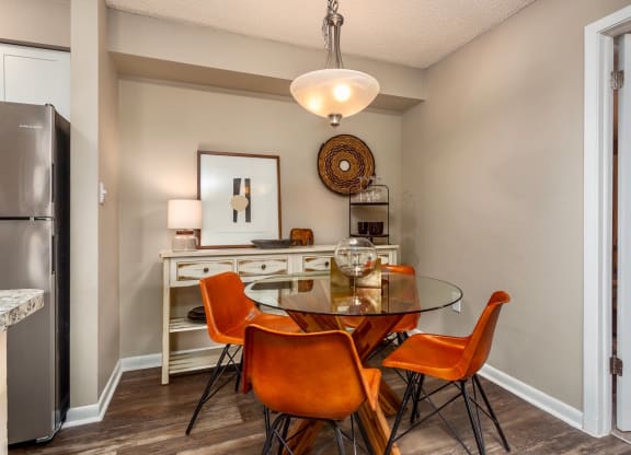 Dining area with a round glass table and four stylish orange chairs. A white sideboard with a modern lamp, framed artwork, and decorative items is placed against the wall. Soft lighting from a pendant light adds warmth to the cozy, contemporary space