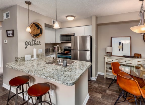 Modern kitchen with a granite countertop, stainless steel appliances, and white cabinetry. The kitchen island features two stools, and the space is decorated with a 'gather' sign on the wall. A dining area with stylish orange chairs and a wooden sideboard is visible in the background, completing the open-concept design
