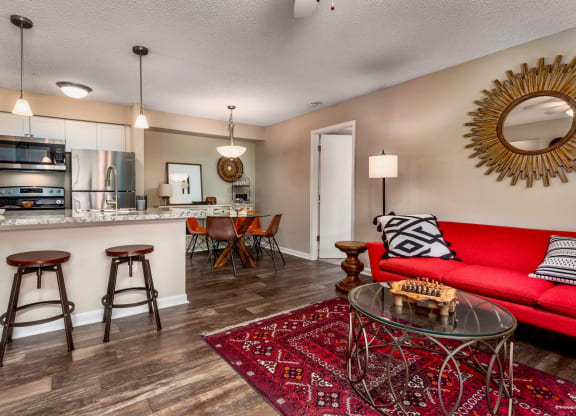 Open-concept living space with a red sofa adorned with black and white pillows. The cozy seating area features a glass coffee table on a vibrant red rug. A kitchen bar with two stools separates the living room from the kitchen, which includes stainless steel appliances and white cabinetry. A dining area with orange chairs and a decorative mirror adds a stylish touch to the room