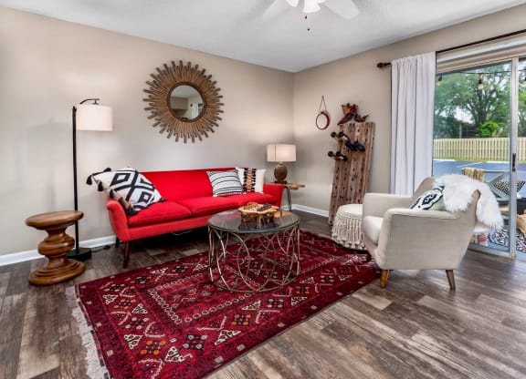Bright living room with a red sofa adorned with black and white pillows. A round glass coffee table sits on a vibrant red rug. The space features a large round mirror on the wall and a comfortable armchair beside a window with white curtains. A unique wooden wine rack and floor lamp complete the modern decor, while natural light floods the room from the open balcony doors