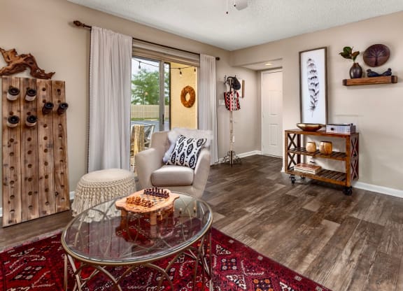 Cozy living room featuring a light-colored armchair and a glass coffee table on a vibrant red rug. Decorative wooden shelves and wall art add a stylish touch. A unique wooden wine rack is placed against the wall, while a large window with white curtains offers a view of the outdoors. A small accent table holds decorative items for a modern, inviting atmosphere