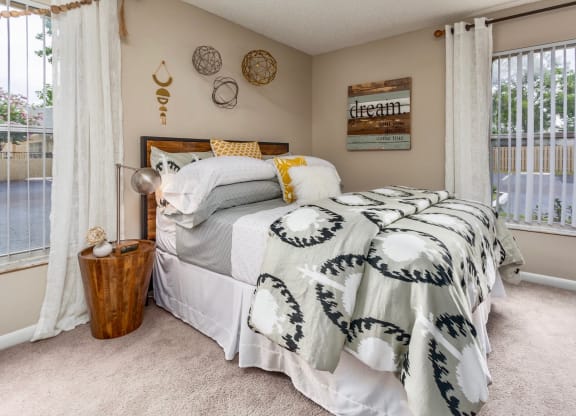 A cozy bedroom with a bed featuring a gray and white patterned blanket, yellow pillow accents, and a framed 'dream' quote on the wall. A wooden side table with a lamp sits beside the bed, and decorative wall pieces enhance the room's ambiance