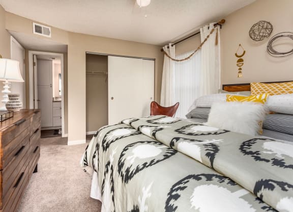 A stylish bedroom featuring a patterned bedspread with gray and yellow accents. A wooden dresser sits beside the bed, and decorative wall art and a hanging light fixture add a cozy touch. A closet and bathroom are visible through an open door
