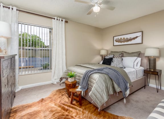 Cozy bedroom featuring a bed with neutral-colored bedding and a throw blanket. The room is decorated with patterned pillows and has a warm, rustic feel with a cowhide rug on the floor. Two side tables with lamps frame the bed, and a wooden dresser sits near the window, which is covered with white curtains. The room also features a decorative feather wall art and a small potted plant