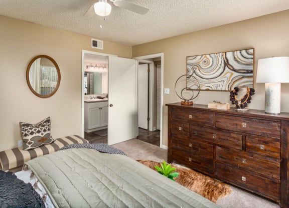 A cozy bedroom with neutral bedding, a wooden dresser, and a large lamp. A round mirror is on the wall, and abstract art hangs above the dresser. The room has minimal decor with a striped bench at the foot of the bed and a view into the bathroom