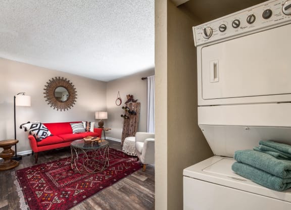 A living room with a red sofa, a patterned rug, and a decorative coffee table. Next to it is a laundry area with a stacked washer and dryer and neatly folded teal towels on top