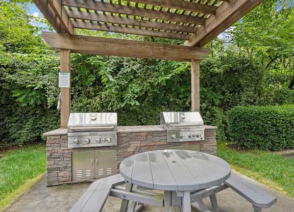 A picnic table is under a pergola next to a stone and metal grill.