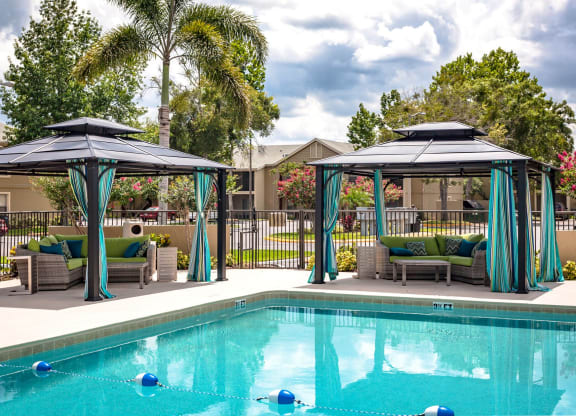 Luxury poolside cabanas with blue and teal curtains, cushioned seating, and a clear blue swimming pool. The area is surrounded by palm trees, lush landscaping, and residential buildings, creating a relaxing outdoor retreat