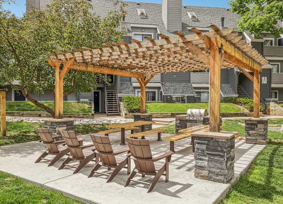 A wooden pergola with chairs and a table is in the foreground of a residential area.