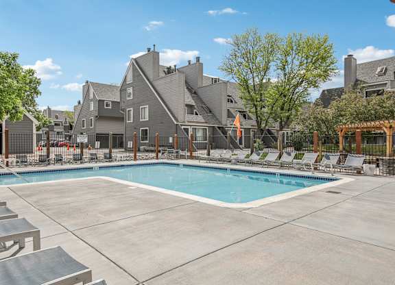 A large swimming pool surrounded by lounge chairs and a fence with a building in the background.
