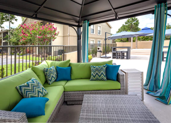 Outdoor cabana lounge with a green sectional sofa, blue accent pillows, and striped teal curtains. The space overlooks a poolside patio with shaded seating and modern outdoor furniture, creating a stylish and relaxing retreat