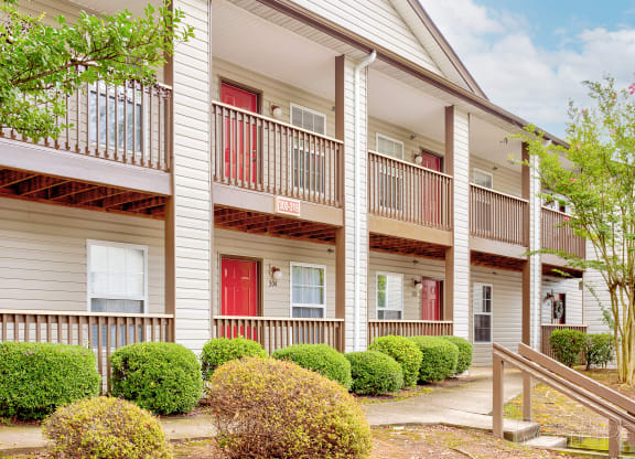 Exterior view of a two-story apartment building featuring beige siding, brown wooden railings, and red front doors. The units have private balconies on the upper level and small porches on the lower level. Lush green shrubs and a well-maintained walkway add to the inviting atmosphere, with a clear blue sky and light clouds in the background