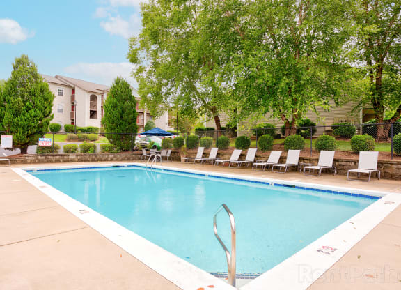 Outdoor swimming pool in a residential community, surrounded by lounge chairs and lush greenery. The pool area is enclosed by a black metal fence with trimmed bushes, and multi-story apartment buildings with balconies are visible in the background. The water is clear and inviting under a bright sky with scattered clouds