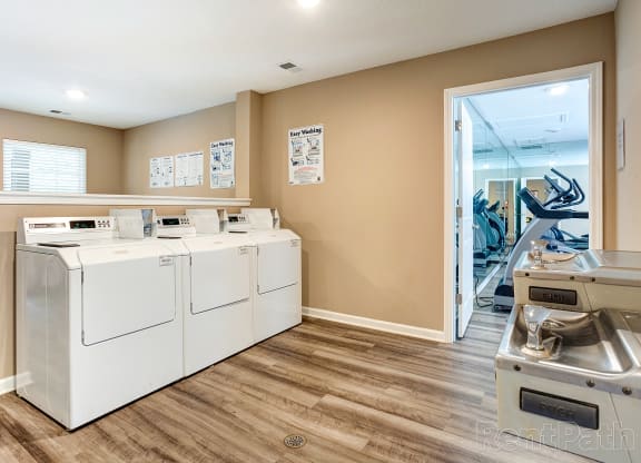 Apartment community laundry room featuring four white top-loading washing machines against beige walls with instructional signs. The space has wood-style flooring, bright overhead lighting, and a window allowing natural light. A doorway leads to an adjacent fitness center