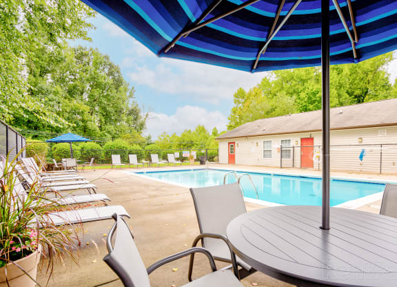 Outdoor swimming pool area with lounge chairs and patio seating under a blue-striped umbrella. The pool is surrounded by lush greenery and a fence, with a small clubhouse or pool house featuring red doors in the background. The water is clear, reflecting the partly cloudy sky, and the area is well-maintained with landscaping and shaded seating options