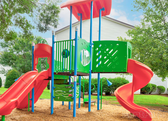 Colorful children's playground with bright red slides, green climbing panels, and blue support poles. The playground features multiple slides, interactive play elements, and a shaded canopy. Surrounded by lush green grass and trees, the playground is set in a residential area with white houses in the background, creating a fun and inviting outdoor space for kids