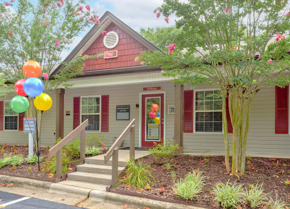 Leasing office with beige siding, red shutters, and a gabled roof. A glass door labeled 'Office' is surrounded by landscaping and colorful balloons near the entrance