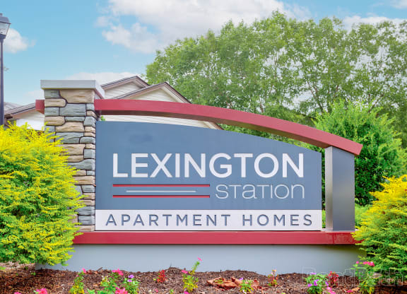 Modern entrance sign for Lexington Station Apartment Homes, featuring a sleek design with stone accents, bold lettering, and a curved red arch, surrounded by lush greenery and flowers