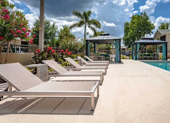 Resort-style pool area with lounge chairs, shaded cabanas with blue curtains, and lush landscaping featuring palm trees and vibrant flowers. The setting offers a relaxing outdoor retreat under a partly cloudy sky